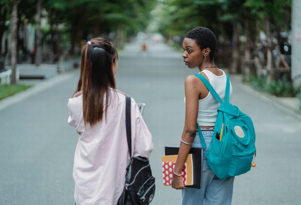 Back view of diverse female students carrying backpacks and supplies for university while talking in park Back view of diverse female students carrying backpacks and supplies for university while talking in park